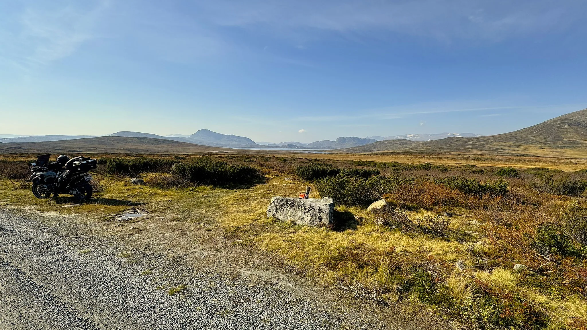 Adventure motorcycle parked beside a gravel road on a high plateau with mountains and a lake in the valley beyond